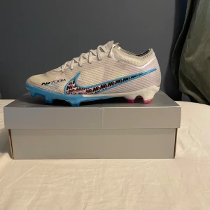 Nike Mercurial fotbollsskor vit/blå/rosa - Nike Mercurial fotbollsskor i vitt med blå och rosa detaljer. Snyggt mönster på sidan och färgglad sula i blått och rosa. Ovandel i syntetmaterial med snörning för bra passform. Perfekta för dig som vill sticka ut på planen.