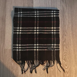Rutig halsduk från Burberrys i ull - Klassisk rutig halsduk från Burberrys of London i 100% lammull. Färgerna är svart, vit och röd med fransar i ändarna. Perfekt accessoar för att lyfta din vinterstil med en tidlös brittisk touch. 