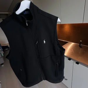 Svart väst från Polo Ralph Lauren - Svart väst från Polo Ralph Lauren med klassisk ståkrage och vit broderad logga på bröstet. Västen har dragkedja framtill, flera fickor med dragkedja och en stilren, sportig look. Nypris kostar denna 1700kr den är knappt använd så säljer den för 900kr 