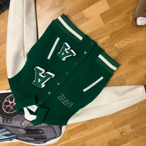 Grön varsityjacka Axel Arigato M - Snygg grön varsityjacka från Axel Arigato med vita ärmar och stora broderade bokstäver på bröstet. Jackan har ribbade muddar, tryckknappar och broderad text på framsidan. Materialmix av ull, polyester och skinnimitation. Perfekt statement-plagg för streetwear.