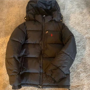 Svart dunjacka från Polo Ralph Lauren - Säljer en svart dunjacka från Polo Ralph Lauren med huva och dragkedja. Jackan har en röd broderad logga på bröstet och flera praktiska fickor. Perfekt för kalla dagar och riktigt skön att ha på sig.
