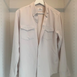 Beige overshirt från Ciszere - Stilren beige overshirt från Ciszere med klassisk krage, två bröstfickor med lock och knappar framtill. Skjortan är långärmad och tillverkad i mjuk bomull, perfekt för lager-på-lager. Enkel och clean look som funkar till många outfits.