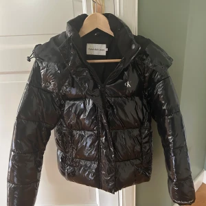 Svart pufferjacka från Calvin Klein Jeans - Säljer en svart, glansig pufferjacka från Calvin Klein Jeans med huva och dragkedja. Jackan har broderad CK-logga på bröstet och Calvin Klein Jeans-tryck på ärmen. Fodrad insida och hög krage för extra värme. Perfekt för kalla dagar. Jätte bra skick inte mycket använd, köpt för 2400