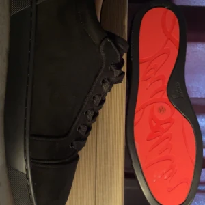 Christian Louboutin svarta sneakers - Svarta sneakers från Christian Louboutin med klassisk röd sula och broderad logga på hälen. Skorna har rund tå, platt sula och snörning. Materialet är mocka och detaljerna är stilrena och lyxiga, perfekt för dig som gillar exklusiv streetstyle.