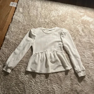 Vit blus med puffärm och volang - Supersöt vit blus med långa puffärmar och volang nedtill. Rund halsringning och lätt rynkad midja ger en feminin siluett. Perfekt att styla med jeans eller kjol för en trendig look.