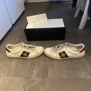 Gucci Ace sneakers med bi-detalj - Gucci Ace sneakers i vitt skinn med klassiska gröna och röda ränder på sidan och broderad guldfärgad bi. Baksidan har metallicröd och metallicgrön häl med Gucci-logga. Snygg och ikonisk modell med snörning och platt sula.