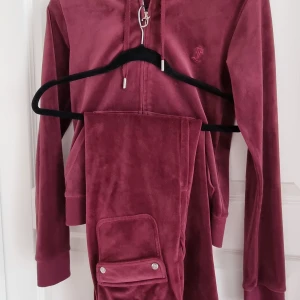 Vinröd velour hoodie från Juicy Couture - Säljer en vinröd hoodie och byxor i mjuk velour från Juicy Couture. Tröjan har huva, halv dragkedja och långa ärmar. Perfekt för en chill och trendig look. Klassisk logga broderad på bröstet och ribbade muddar vid ärmslut. Xs på tröjan och xxs på byxorna. 