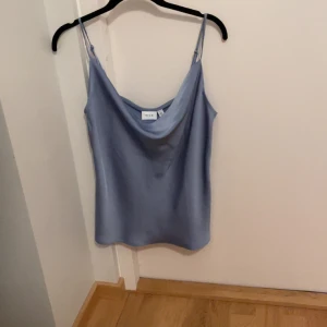 Ljusblå linnetopp från Vila - Säljer en ljusblå linnetopp från Vila med tunna axelband och snygg draperad urringning. Toppen har en slät, glansig yta som ger en lyxig känsla och passar perfekt till jeans eller kjol. Enkel och stilren design som är lätt att matcha.