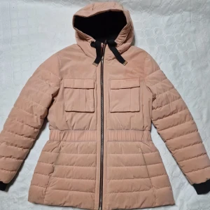 Beige pufferjacka från Moose Knuckles - Snygg beige pufferjacka från Moose Knuckles med huva, svarta detaljer och två stora fickor framtill. Jackan har dragkedja, quiltad design och elastisk midja för en schysst passform. Perfekt för kalla dagar och riktigt stilren look. Äkta. 