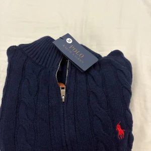 Quarter zip Mörkblå stickad tröja Polo Ralph Lauren - Kabelstickad mörkblå tröja från Polo Ralph Lauren med halv dragkedja och klassisk röd logga på bröstet. Tröjan har ribbade muddar och hög krage, perfekt för lager-på-lager. Snygg och tidlös design som funkar året runt.