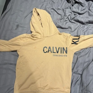 Beige hoodie från Calvin Klein Jeans - Snygg beige hoodie från Calvin Klein Jeans med svart tryck på bröstet och ärmen. Tröjan har huva och långa ärmar, perfekt för chill dagar. Materialet är mjukt och skönt bomullstyg. Stort CALVIN-tryck framtill och JEANS-tryck på ryggen ger en cool streetkänsla.