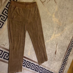 Bruna mjukisbyxor i sammet - Bruna juicy couture mjukisbyxor i mjuk sammet med resårmidja och snörning. Byxorna har två fickor fram och två bakfickor med lock och knappar. Perfekta för en chill och avslappnad stil.   Använd byxor 