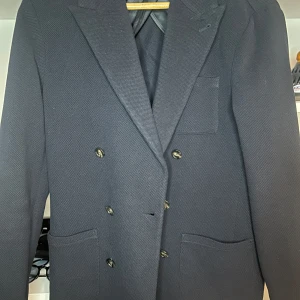 Mörkblå dubbelknäppt kavaj John Henric - Snygg mörkblå dubbelknäppt kavaj från John Henric. Klassisk modell med breda slag, fyra fickor och sex knappar framtill. Tyg i strukturerad ullmix och helfodrad insida. Perfekt för dig som vill ha en stilren och tidlös look.