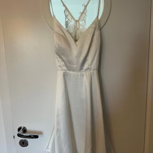 Vit kort klänning Moments New York - Superfin vit kort klänning från Moments New York med smala axelband, v-ringning och spetsdetaljer i ryggen. Klänningen har en lätt glansig satinlook och dragkedja i sidan. Perfekt för dig som vill ha en elegant och stilren look.