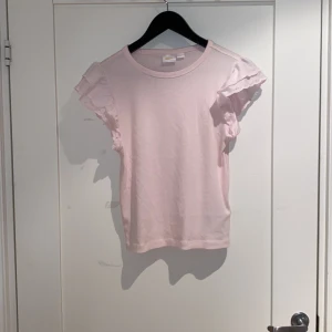 Ljusrosa topp med volangärmar Gina Tricot - Supersöt ljusrosa topp från Gina Tricot med korta volangärmar och rund halsringning. Tillverkad i mjuk bomull, perfekt för dig som gillar en feminin och enkel stil. Passar till jeans eller kjol för en chill look. Säljer tröjan eftersom att den inte kommer till användning.🩷