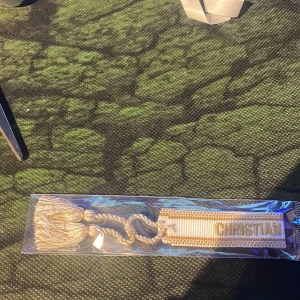 Beige Christian Dior J'ADIOR armband - Snyggt beige armband från Christian Dior med broderad text 'CHRISTIAN DIOR J'ADIOR' i guld. Armbandet har flätade band och två tofsar i ändarna för en cool och lyxig vibe. Perfekt accessoar för att lyfta din outfit.