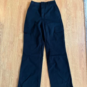 Svarta cargopants från A DSGN XXS - Svarta cargopants från A DSGN i storlek XXS. Byxorna har raka ben, hög midja och stora fickor på sidorna. Tillverkade i ett slitstarkt bomullstyg med bälteshällor och knappstängning. Perfekta för en chill och streetig look.