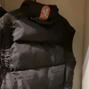 Svart dunväst med fuskpäls-krage, Parajumper - Snygg svart dunväst med mjuk fuskpäls på kragen och dragkedja framtill. Västen har två stora fickor med knappar och dragkedjor, samt quiltad design som ger en cool look. Perfekt för lager-på-lager under kyliga dagar. Säljer denna för så billigt för att en av fickorna är öppen och går inte stänga då zipen har gått bort men resten ser bra ut.