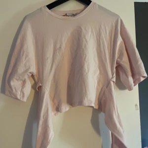 Ljusrosa croppad topp med knyt - Supersöt ljusrosa croppad topp med korta ärmar och långa band som knyts framtill i en stor rosett. Toppen har en enkel och clean design i mjuk bomull, perfekt för en chill och trendig look. (FLÄCK SOM SYNs PÅ BILD 1&4 går nog att få bort) 