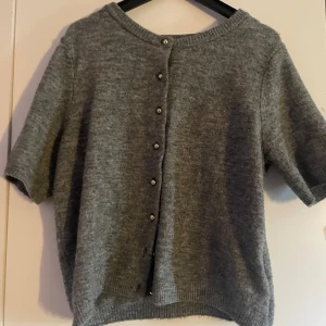 Grå kofta med knappar från H&M - Mysig grå kofta från H&M med korta ärmar och snygga knappar längs hela framsidan . Koftan är stickad och har en rund halsringning samt ribbade muddar vid ärmslut och nederkant. Perfekt för lager-på-lager-stil under kyliga dagar.