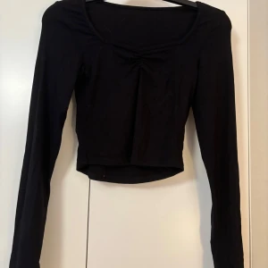 Svart croppad topp från Dazy - Svart långärmad topp från Dazy med croppad passform och lätt rynkdetalj vid bysten. Toppen har en rundad halsringning och är tillverkad i mjuk bomullsmix. Perfekt för en stilren och enkel look.