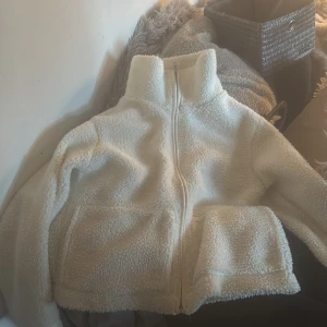 Vit teddyjacka från 157 - Mysig vit teddyjacka från 157 med hög krage och dragkedja framtill. Jackan är croppad och har en oversized passform, perfekt för kyliga dagar. Tillverkad i mjukt syntetmaterial som känns fluffigt och varmt.