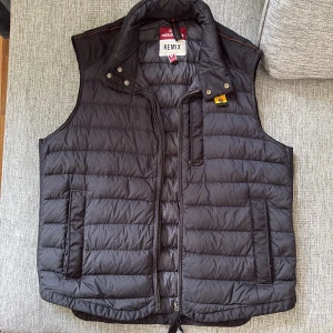 Svart dunväst från Parajumpers Remix - Svart dunväst från Parajumpers Remix med quiltad design och hög krage. Västen har dragkedja framtill, två sidofickor och snygga detaljer vid halsen. Perfekt för lager-på-lager och har en stilren look med diskret logga på insidan. Qr kod för äkthet finns på insidan. Bild med väst på samt mått kan skickas vid intresse.