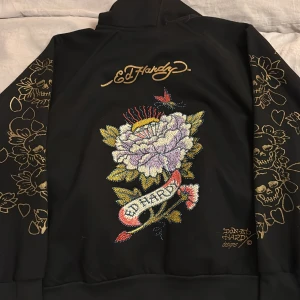 Svart Ed Hardy hoodie med blommor  - Svart hoodie från Ed Hardy med ikoniska guldiga blom- och dödskalletryck på ärmarna och stor broderad blomma i lila, vitt och gult på ryggen. Dragkedja framtill, huva och coola detaljer som Ed Hardy-logga på bröstet. Perfekt statement-plagg för dig som gillar streetstyle.