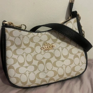 C axelväska  - Snygg axelväska i beige och vitt med klassiskt monogrammönster och guldfärgade detaljer. Väskan har svart axelrem och dragkedja upptill. Perfekt för dig som vill ha en stilren och trendig accessoar.