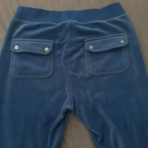 Blå velourbyxor Juicy Couture - Mysiga blå mjukisbyxor från Juicy Couture i velour med två stora fickor bak med silverfärgade knappar och broderad JC-logga framtill. Perfekta för chill dagar och har en klassisk, avslappnad look.
