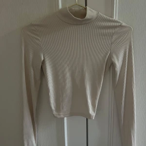 Beige ribbad polotopp med twistad detalj - Säljer en beige ribbad polotopp med lång ärm och croppad passform. Toppen har en snygg twistad omlottdetalj framtill som skapar en öppen rygg. Perfekt för dig som gillar trendiga och unika plagg med det lilla extra.