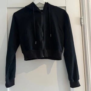 Svart croppad hoodie med dragkedja - Svart croppad hoodie med dragkedja framtill och huva med snörning. Tröjan har långa ärmar och bred mudd vid midjan och ärmsluten. Perfekt för en chill och avslappnad stil.