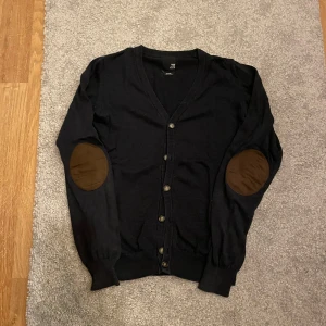 Navy Cardigan  - Mörkblå kofta med v-ringning och knäppning framtill. Långa ärmar och snygga bruna armbågslappar som ger en klassisk touch. Perfekt för lager-på-lager under hösten eller vintern.