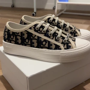 Christian Dior sneakers  - Snygga sneakers från Christian Dior i beige och marinblått monogramtyg. Skorna har vit platt sula, rund tå och snörning framtill. Tydlig Dior-logga på sidan och på snörningen. Perfekta för dig som vill sticka ut med exklusiv streetstyle.