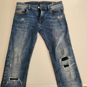 Blå slitna jeans från Diesel - Säljer ett par blå jeans från Diesel med snygga slitningar och hål på benen. Jeansen har klassisk femficksdesign, normal passform och tydliga slitdetaljer för en cool look. Materialet är robust denim i bomull. Perfekta för dig som gillar en avslappnad och trendig stil.