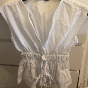 Vit omlottopp med knyt i midjan - Säljer en vit ärmlös omlottopp med djup v-ringning och knytband i midjan. Toppen har en luftig passform med rynkade detaljer och volangliknande nederkant. Perfekt för varma dagar och enkel att styla till olika looks.