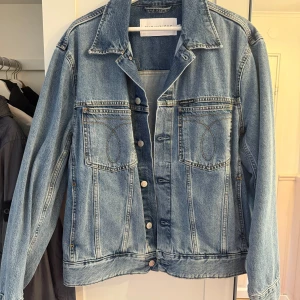 Ljusblå jeansjacka från Calvin Klein - Snygg ljusblå jeansjacka från Calvin Klein Jeans med klassisk passform. Jackan har två bröstfickor med lock och knappar framtill, samt diskret logotyp på bröstet och nertill på ryggen. Perfekt oversized vibe och tillverkad i slitstarkt denim.
