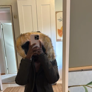 Svart parkasjacka med fuskpäls från Subprime - Säljer en mörkgrön parkasjacka från Subprime med röd insida och stor huva med fuskpäls. Jackan har dragkedja och knappar framtill, samt en patch på ärmen. Perfekt för kalla dagar med sin långa modell och varma foder.