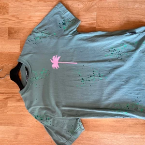 Grön Palm Angels t-shirt med tryck - Snygg grön t-shirt från Palm Angels med rosa palmtryck framtill och rosa text på ryggen. T-shirten har gröna färgstänk över hela plagget och är tillverkad i 100% bomull. Klassisk passform med rund hals och korta ärmar.