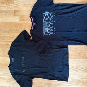 Svarta t-shirts Hugo Boss & Armani - Två svarta t-shirts, en från Hugo Boss med stor logga och mönstrad detalj framtill, och en från Emporio Armani med diskret logotyp på bröstet. Båda har rund hals och korta ärmar. Perfekta för en clean och stilren look.