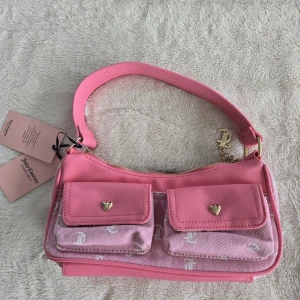 Rosa axelväska från Juicy Couture - Superfin rosa axelväska från Juicy Couture med två fickor framtill som har hjärtformade knappar. Väskan har både släta och mönstrade partier med JC-logga, guldfärgade detaljer och dragkedja upptill. Insidan är fodrad med Juicy Couture-tryck. Perfekt för dig som gillar en söt och trendig look.