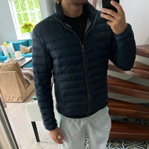 Colmar jacka - Säljer en mörkblå pufferjacka från Colmar med horisontella sömmar, hög krage och två dragkedjeförsedda fickor. Jackan har Colmar-logga på ärmen och insidan är ljusgrå. Perfekt för kyliga dagar och riktigt snygg till streetwear. Personen på bilden är 180cm. Storlek 50, sitter som S/M