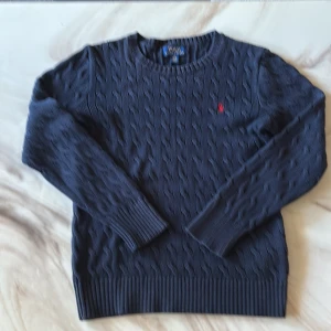 Mörkblå kabelstickad tröja Polo Ralph Lauren - Kabelstickad mörkblå tröja från Polo Ralph Lauren med klassisk röd broderad logga på bröstet. Tröjan har rund halsringning, långa ärmar och ribbade muddar vid ärmslut och nederkant. Perfekt för dig som gillar stilren och tidlös design.