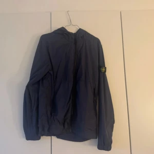 Mörkblå vindjacka från Stone Island - Snygg mörkblå vindjacka från Stone Island Junior med huva och klassisk logga på ärmen. Jackan är tillverkad i lätt polyester och har dragkedja framtill. Perfekt för dig som vill ha en stilren och sportig look. Den är junior storlek men passar storlek S, kvitto finns också