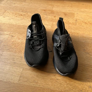 Svarta sneakers med guldtext,  - Svarta sneakers i storlek 35 med cool guldtext på sidan och ovandel i mesh. Skorna har snörning, platt sula och detaljer i syntetmaterial. Perfekta för dig som gillar sportig och modern stil.