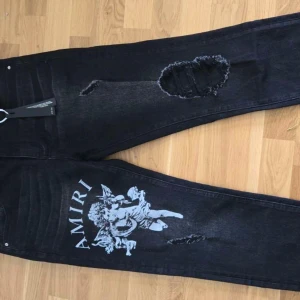 Svarta Amiri jeans med ängeltryck - Svarta jeans från Amiri med stort vitt ängeltryck och logga på ena benet. Snygga slitningar och hål på benen ger en edgy look. Klassisk femficksmodell i jeansmaterial med raka ben och metallmärke bak på fickan.