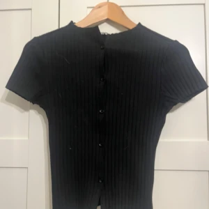 Svart ribbad topp från Gina Tricot - Svart ribbad topp från Gina Tricot med korta ärmar och knappar längs ryggen. Toppen har en tight passform och är tillverkad i ett stretchigt material som sitter snyggt på kroppen. Perfekt att styla med jeans eller kjol.