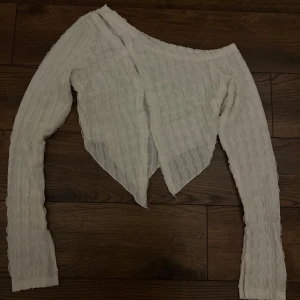Vit offshoulder topp med vågig struktur - Säljer en vit långärmad topp med offshoulder-skärning och vågig struktur i tyget. Toppen har en spetsig nederkant framtill och är tillverkad i ett lätt, transparent material som ger en cool och trendig look.