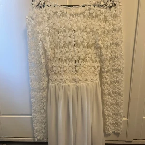 Vit spetsklänning från DM Retro - Superfin vit klänning från DM Retro med långärmade spetsärmar och blommigt spetsmönster upptill. Kjolen är slät och faller snyggt. Perfekt för dig som gillar romantisk och feminin stil. Dragkedja i bak för enkel påklädning.