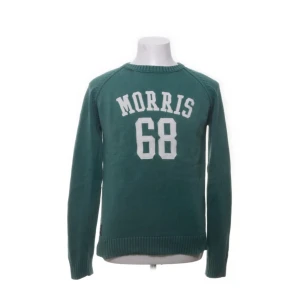 Grön stickad tröja Morris med tryck - Grön stickad tröja från Morris med vitt tryck 'Morris 68' på bröstet och svarta armbågslappar. Klassisk rund halsringning och ribbade muddar. Perfekt för dig som gillar sportig och preppy stil.